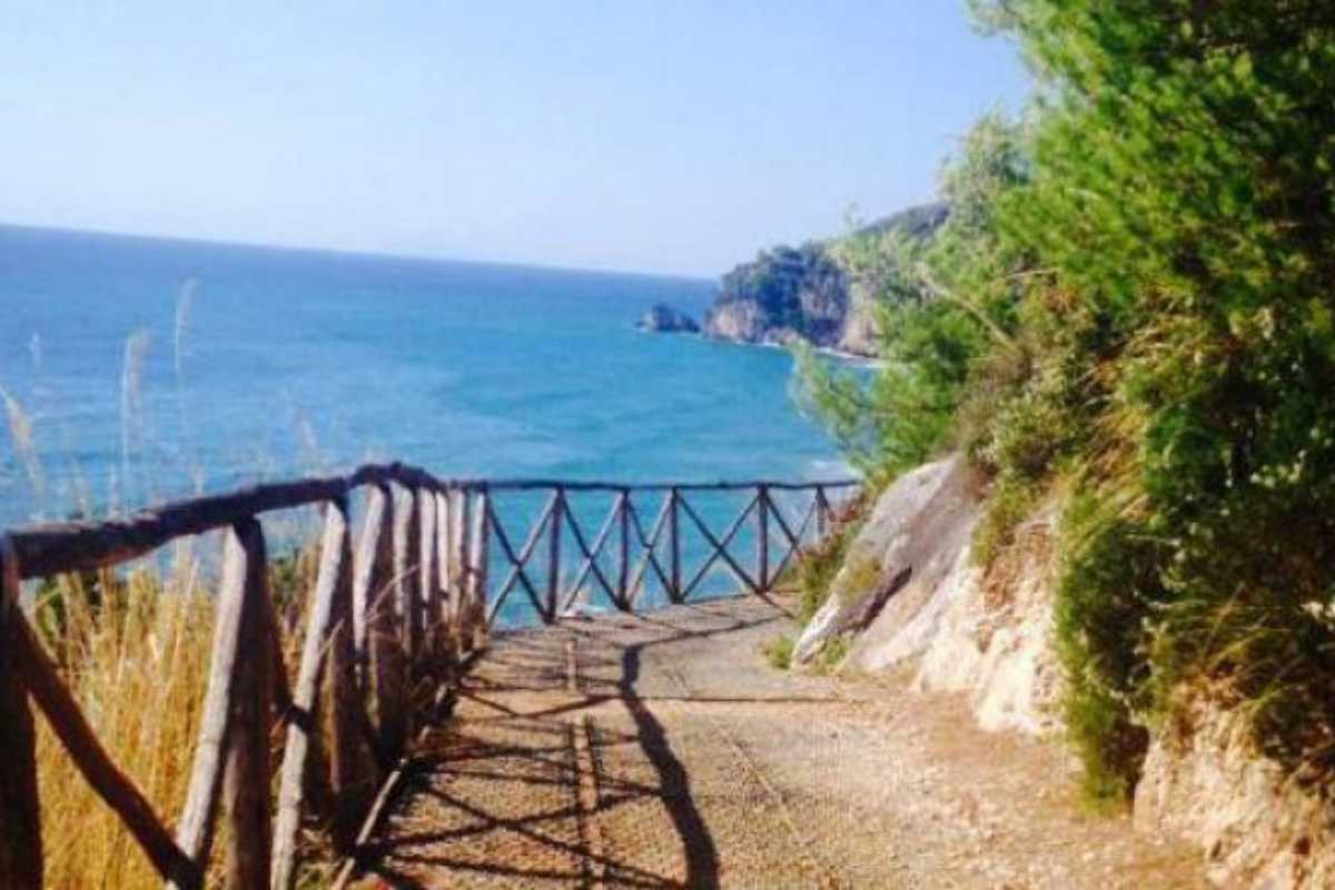Vista panoramica dal sentiero costiero sopra Sperlonga, con la città bianca sullo sfondo e il mare azzurro. Luce dorata del mattino, atmosfera primaverile.